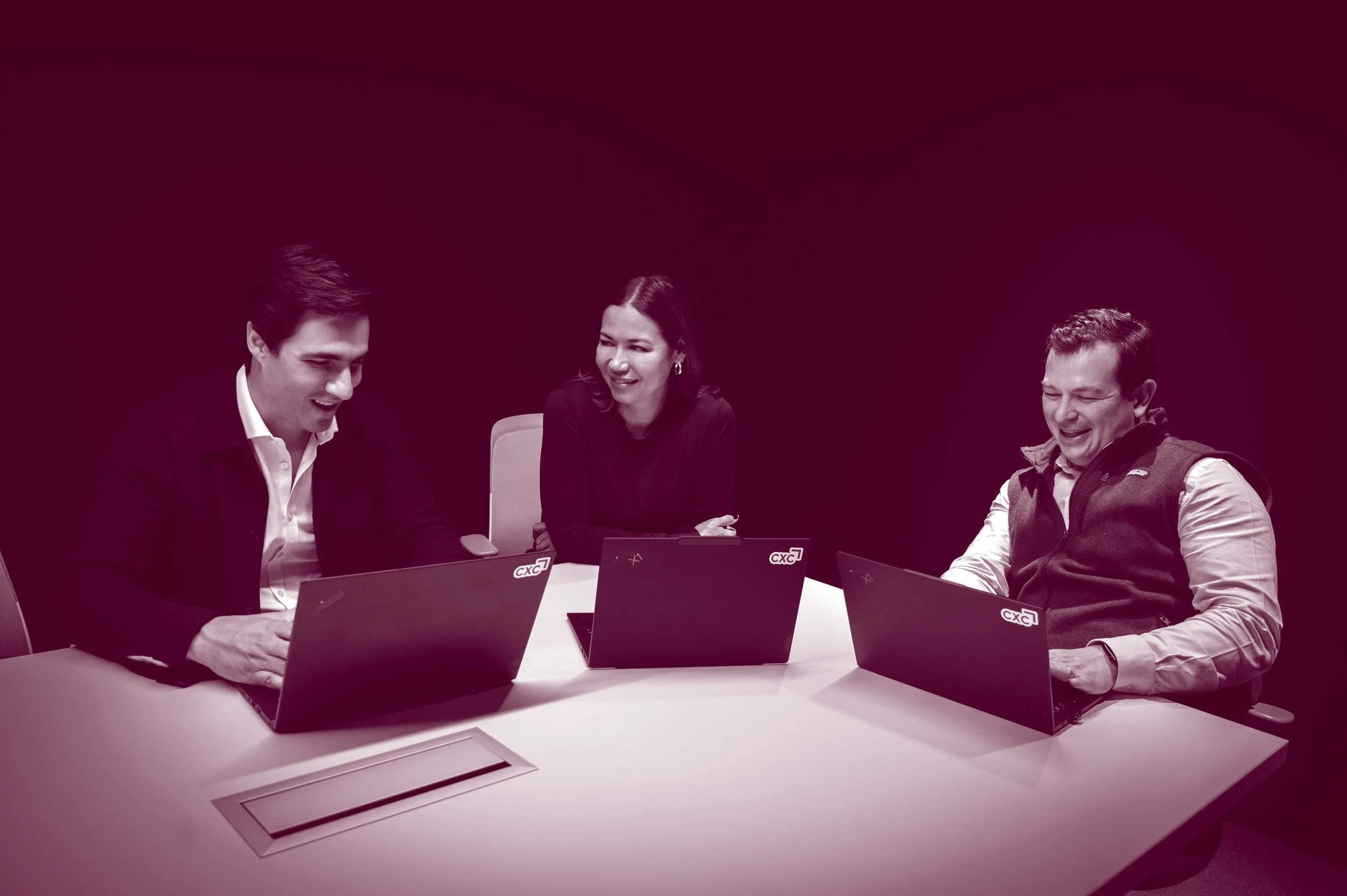 Three colleagues laughing while working together on laptops in dark room.